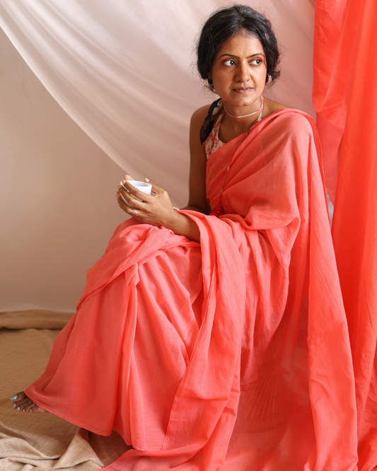A woman sits gracefully draped in Dawn, a handwoven lightweight cotton saree in a soft coral hue. The saree’s airy texture and fluid drape capture the gentle warmth of early morning light, blending traditional craftsmanship with everyday elegance — perfect for slow mornings and timeless, effortless style.