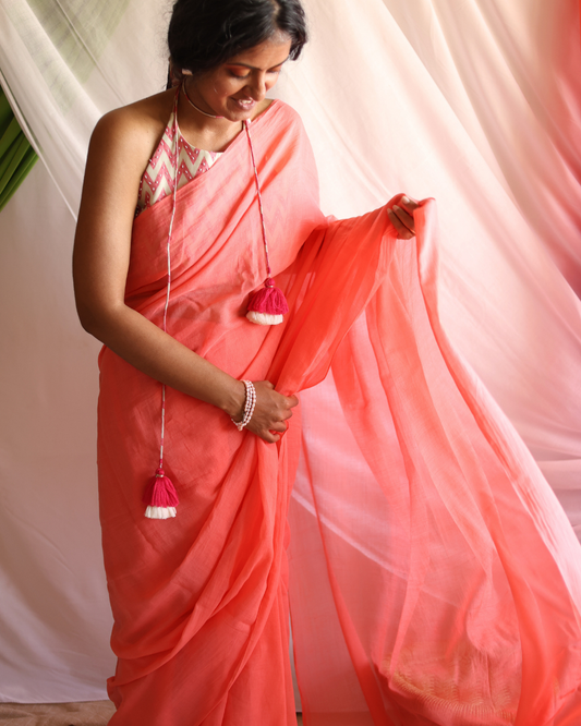A woman smiles softly while draped in Dawn, a handwoven coral cotton saree known for its feather-light texture and graceful drape. The saree’s soft, sunrise-inspired hue and flowing silhouette make it perfect for everyday wear or special occasions, celebrating handcrafted elegance and sustainable fashion.