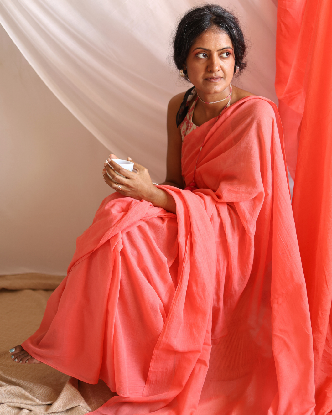 A woman sits gracefully draped in Dawn, a handwoven lightweight cotton saree in a soft coral hue. The saree’s airy texture and fluid drape capture the gentle warmth of early morning light, blending traditional craftsmanship with everyday elegance — perfect for slow mornings and timeless, effortless style.