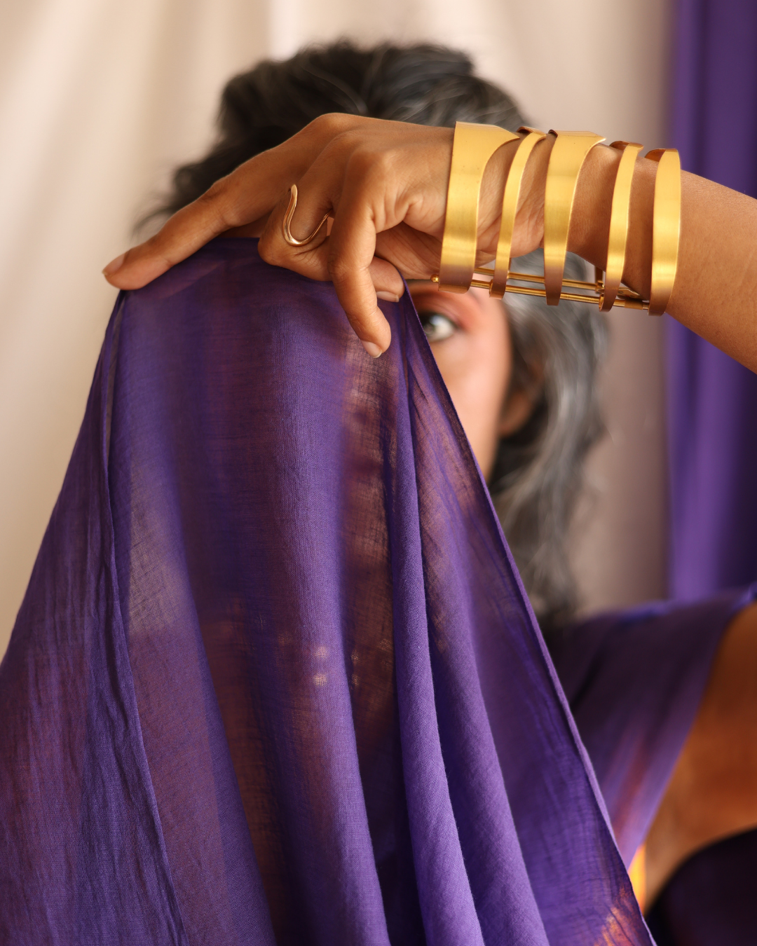 Close-up of the GALAXY solid purple handloom saree held gently by a woman wearing bold gold jewellery. This handcrafted cotton saree is airy, minimal, and elegant — a slow-made piece that blends traditional weaving with timeless, versatile style.
