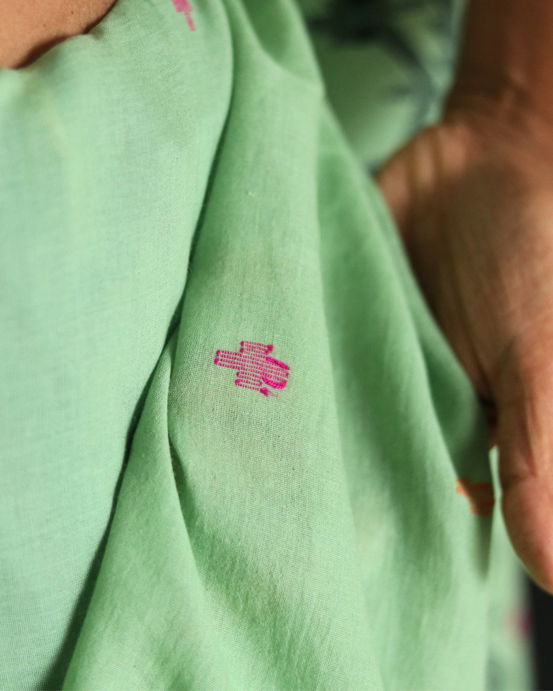 Close-up of a soft green handwoven Bengal cotton saree showcasing delicate pink woven motifs. Highlights the craftsmanship and precision of traditional weaving techniques that make the fabric breathable, lightweight, and perfect for everyday elegance.