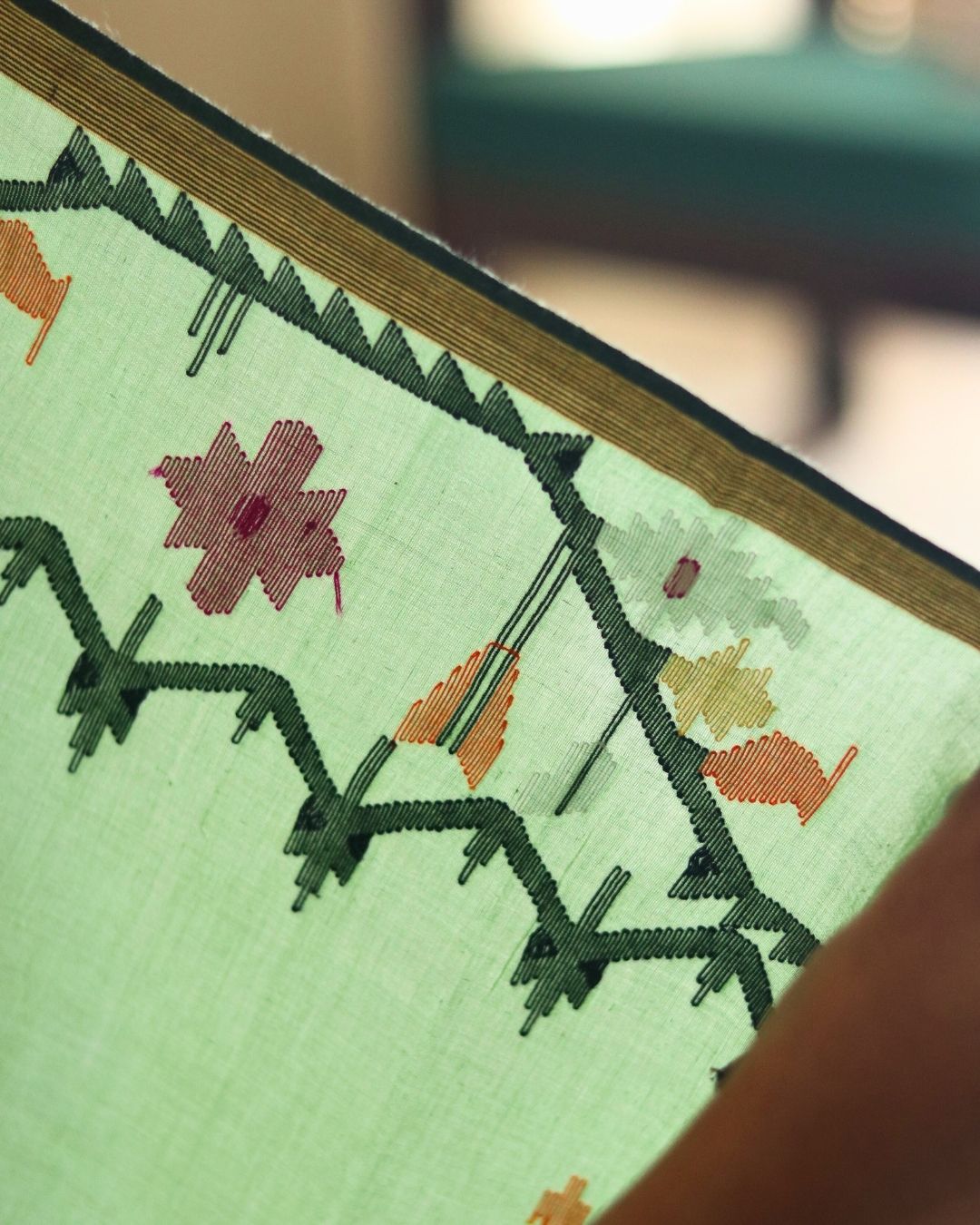 Close-up of the handwoven floral motifs on a light green Bengal cotton saree, showcasing intricate weaving patterns and vibrant threadwork in pink, orange, and white. This detailed shot highlights the craftsmanship and traditional artistry behind the saree’s elegant border design.
