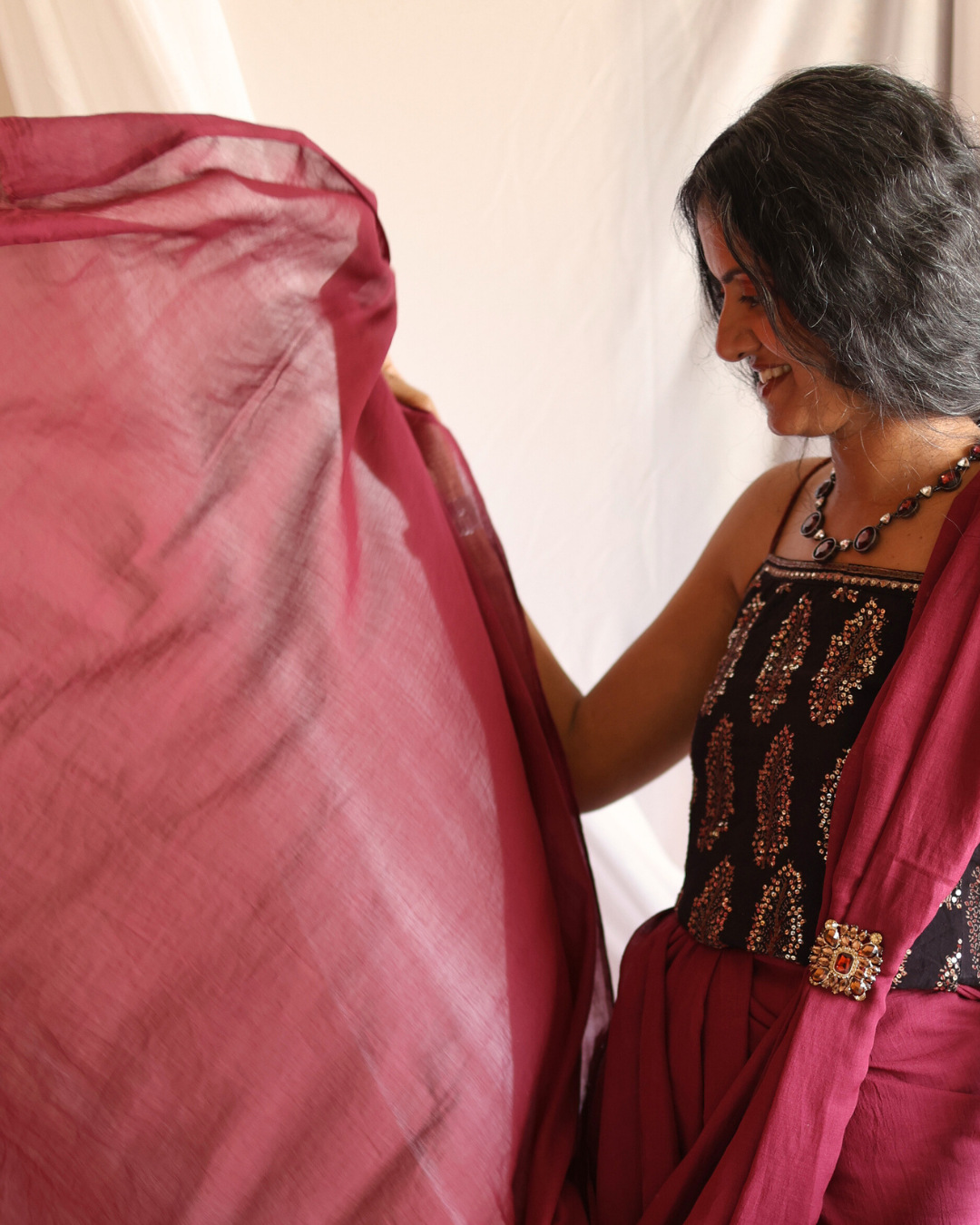 A woman draped in the POMEGRANATE pure Bengal cotton saree, showcasing its rich red color and lightweight, handwoven texture. The image highlights the airy pallu as she holds it up, paired with an embroidered black blouse and handcrafted jewelry — a timeless blend of traditional craftsmanship and modern style.