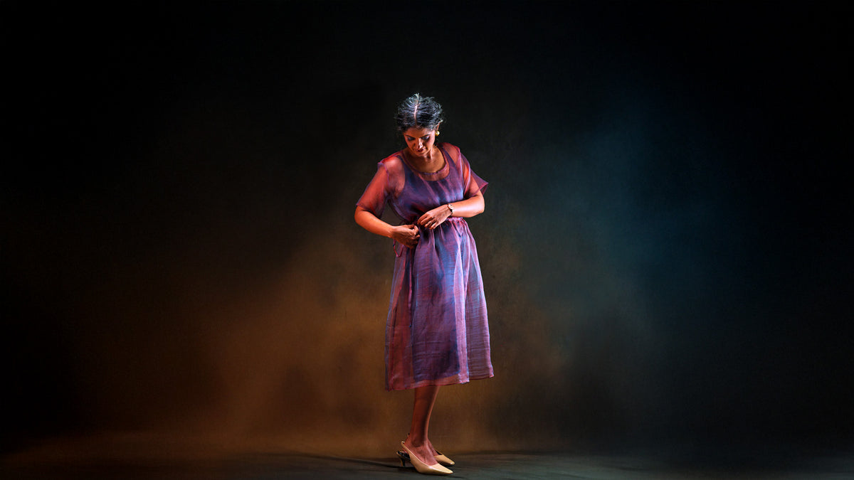 A woman in a sheer silk red and blue dress stands on a dark stage under dramatic spotlighting.