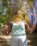 Woman in a light green top and white pants standing outdoors with a blurred natural background