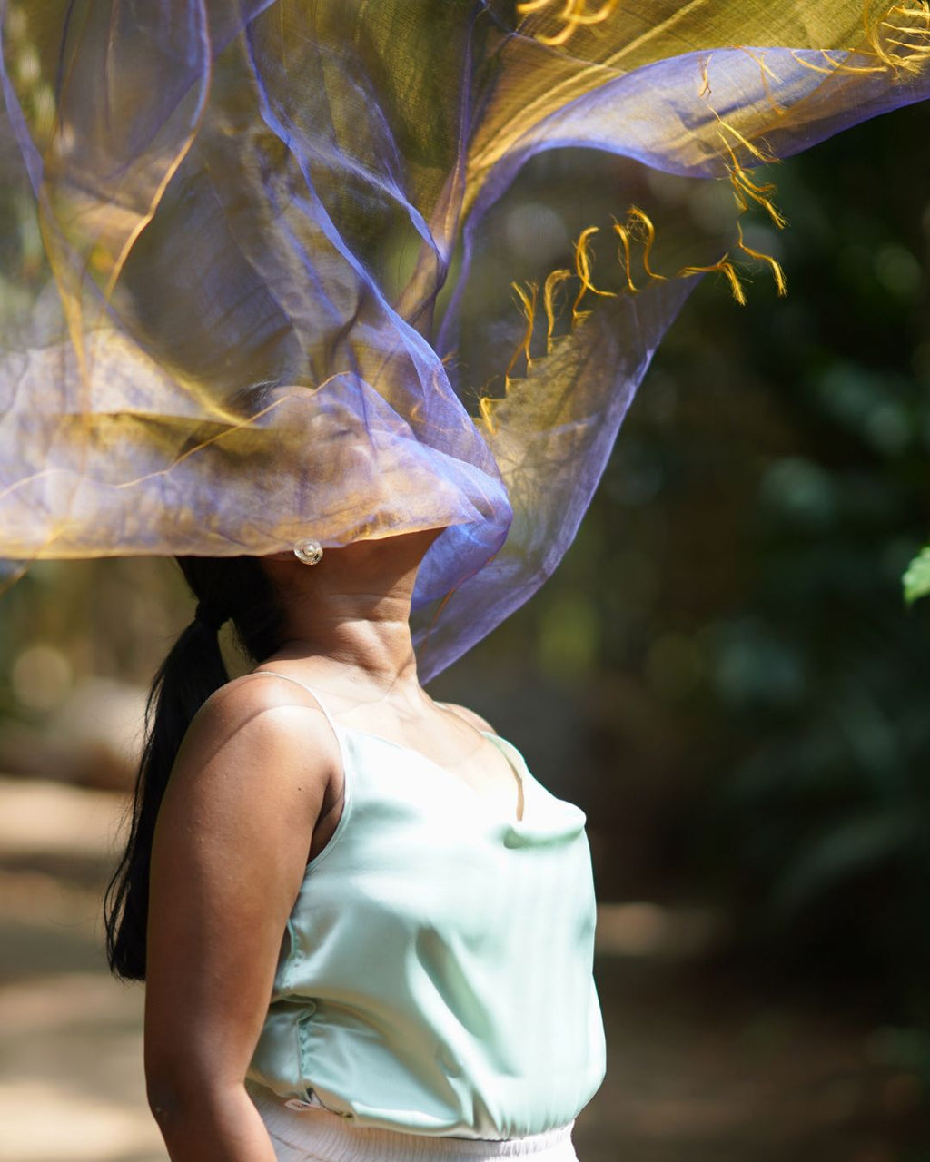Woman wearing a light blue sleeveless top with a sheer yellow and purple fabric draped over her head in a natural setting.
