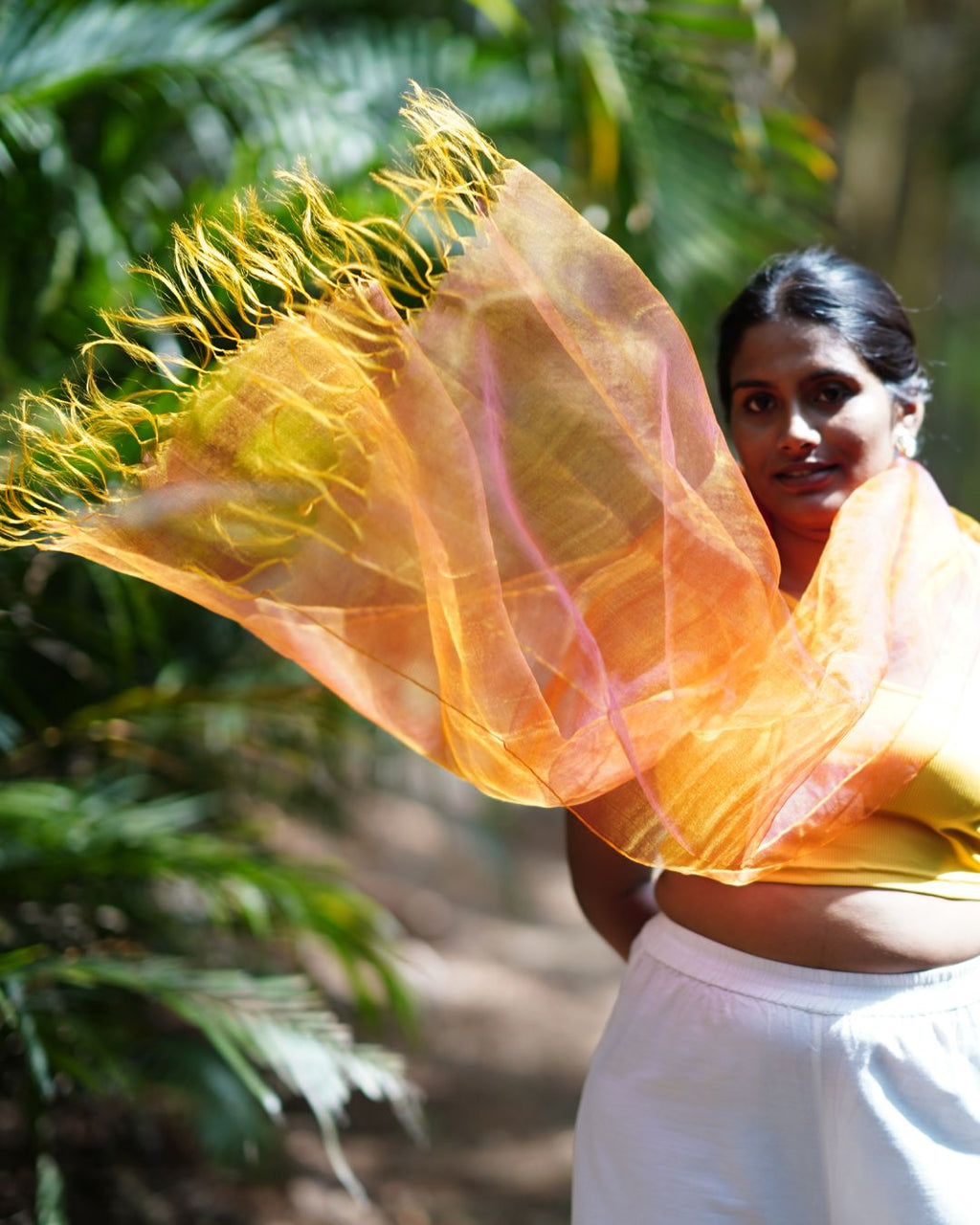 Woman holding a yellow and orange scarf with green foliage in the background