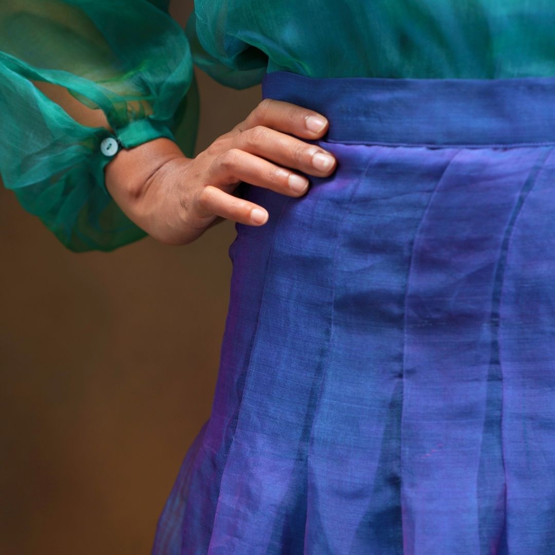 Close-up of a person wearing a pleated skirt in a blue purple silk, with a sheer teal green blouse and one hand resting on the waistband.