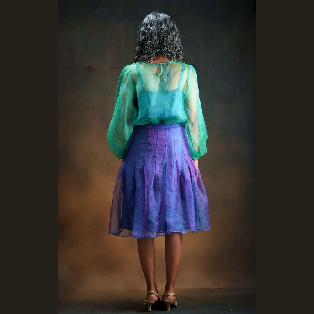 A woman viewed from behind wearing a sheer long-sleeve green top and a pleated silk knee length skirt in purple and blue standing against a dark brown studio backdrop.