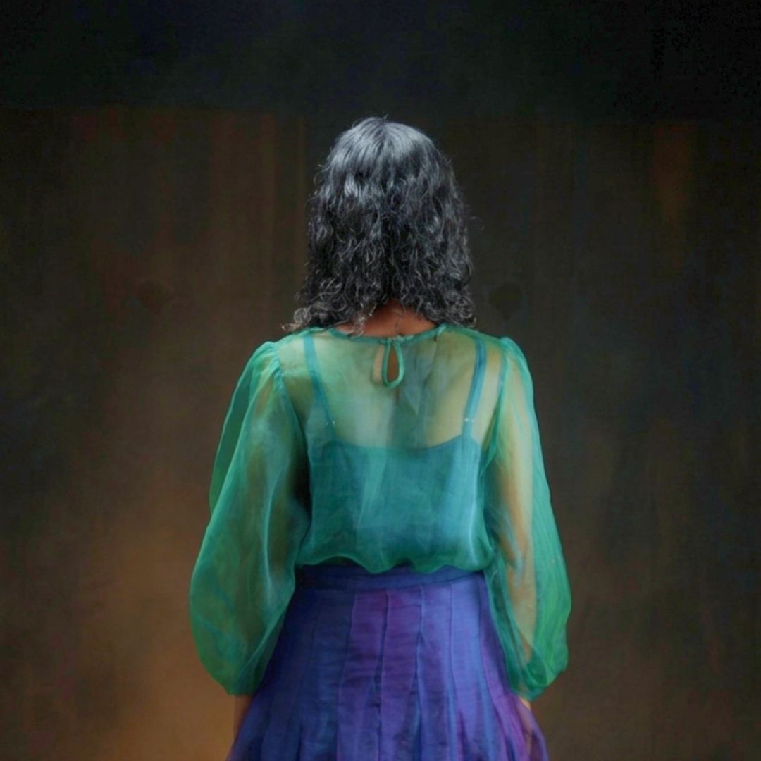 A person viewed from behind wearing a sheer teal green long-sleeve top over a teal camisole and a purple skirt, standing against a dark brown textured background.