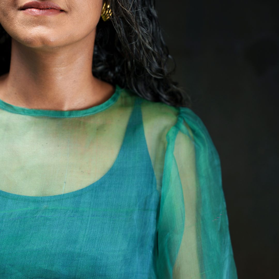 A close-up of a woman wearing a sheer teal green long-sleeve top with a solid teal underlayer, set against a dark background.