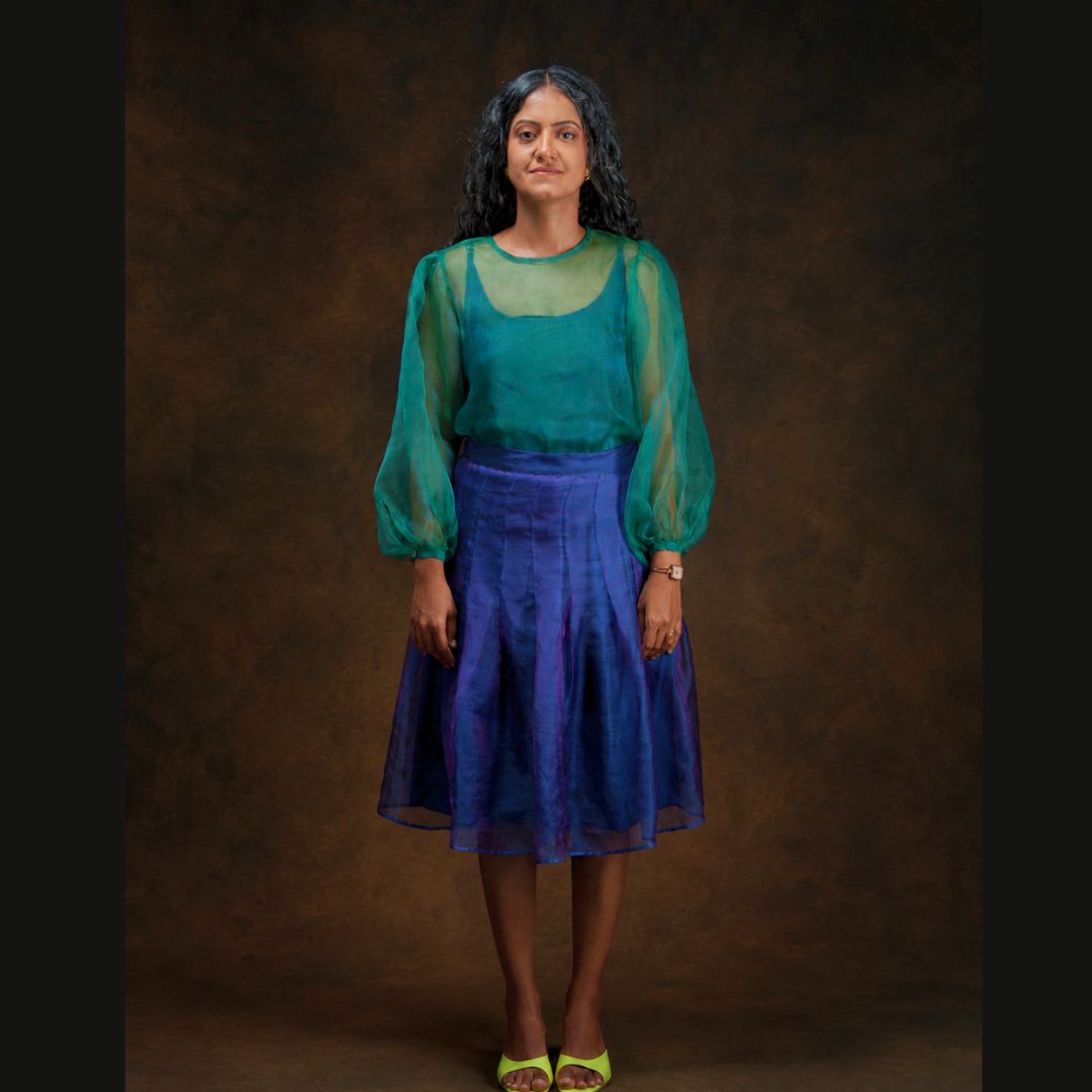 A woman stands against a dark brown studio backdrop wearing a sheer teal green long-sleeve top and a blue-purple pleated midi skirt, with neon yellow sandals.