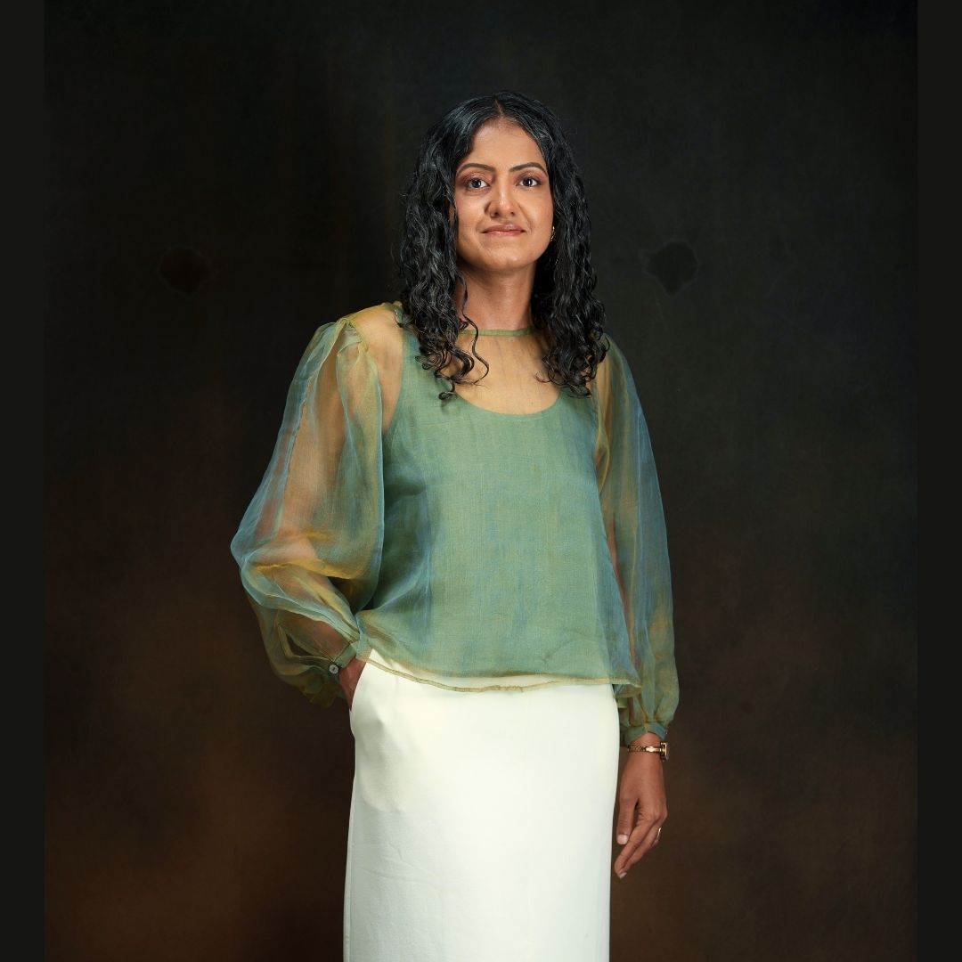 A woman with dark curly hair stands against a dark brown studio background, wearing a gold andsage green sheer long-sleeve blouse with a white skirt.