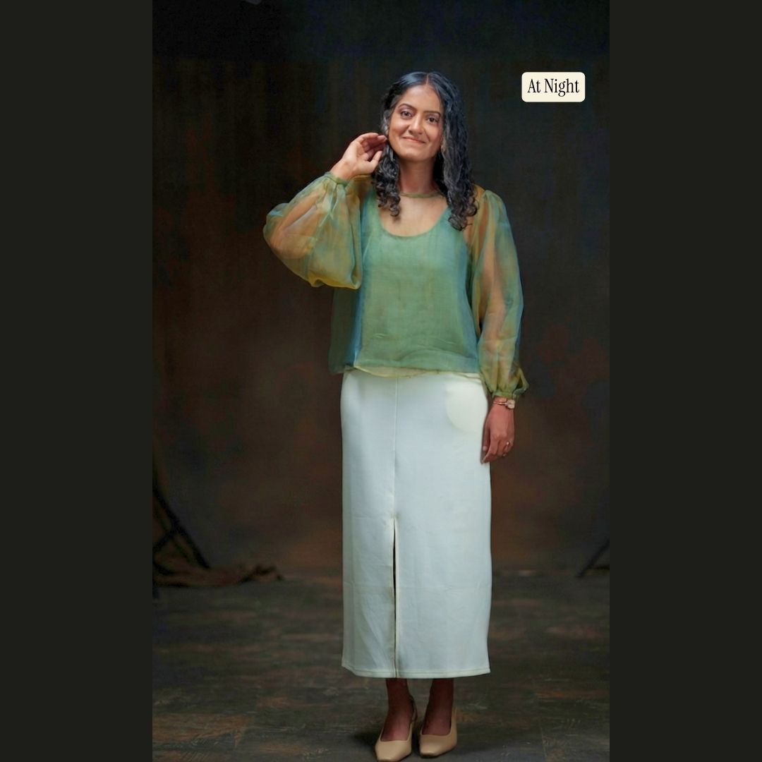 A woman modeling a two-piece outfit consisting of a sage green sleeveless tank top and a matching sheer gold olive green balloon sleeve top, paired with a cream midi skirt with a front slit and beige heels. The image has a dark studio background with a 'at night' label in the upper right.