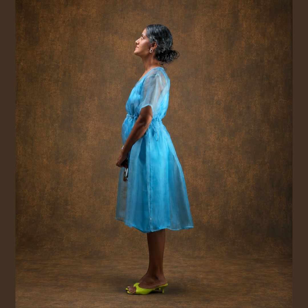 A woman in profile wearing a sheer light blue midi dress with short sleeves and lime green heeled sandals, standing against a textured brown studio backdrop.