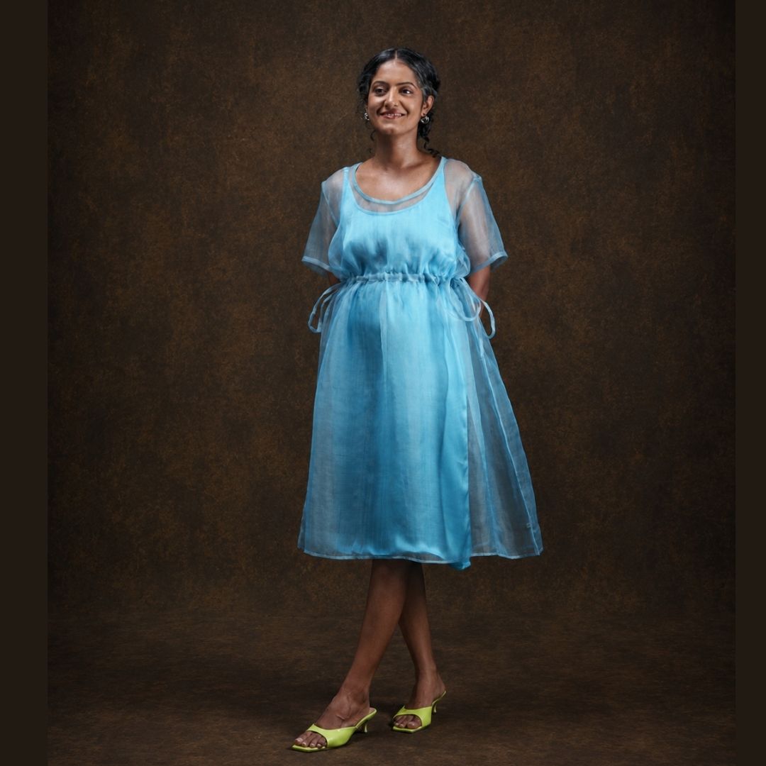 A woman modeling a light blue midi silk dress with short sleeves and a drawstring waist, standing against a textured brown studio backdrop.