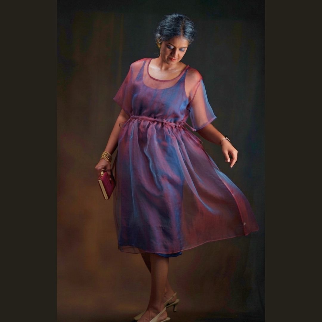 A woman wearing a sheer, flowy silk midi dress with a blue and red dual tone holding a small burgundy clutch against a dark brown studio background.