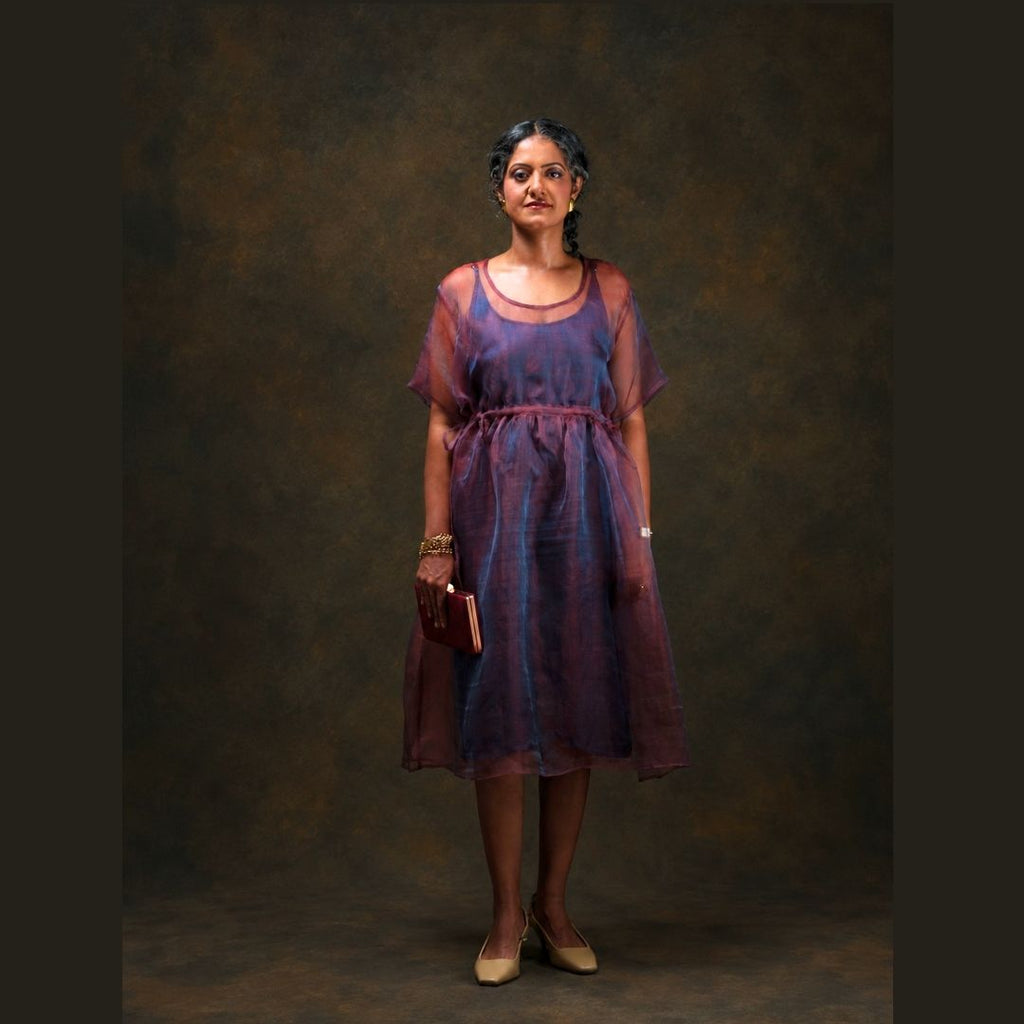 A woman stands against a dark brown studio backdrop wearing a sheer red and blue silk midi dress with a dark blue inner dress. She is holding a small dark red clutch and wearing beige heels.