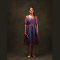 A woman stands against a dark brown studio backdrop wearing a sheer red and blue silk midi dress with a dark blue inner dress. She is holding a small dark red clutch and wearing beige heels.