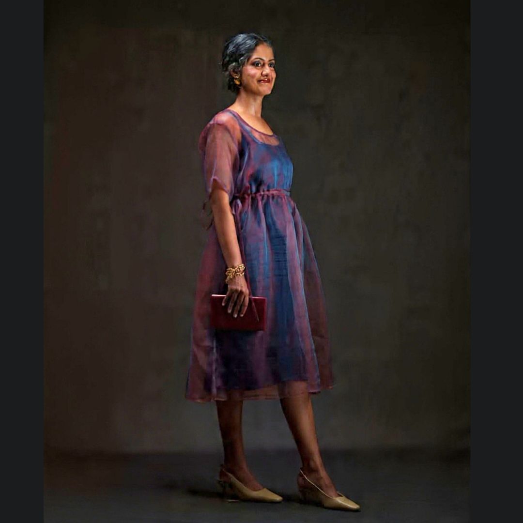 A woman modeling a sheer red and blue silk midi dress with a blue inner slip, paired with beige heels and holding a dark red clutch, set against a dark textured studio backdrop.